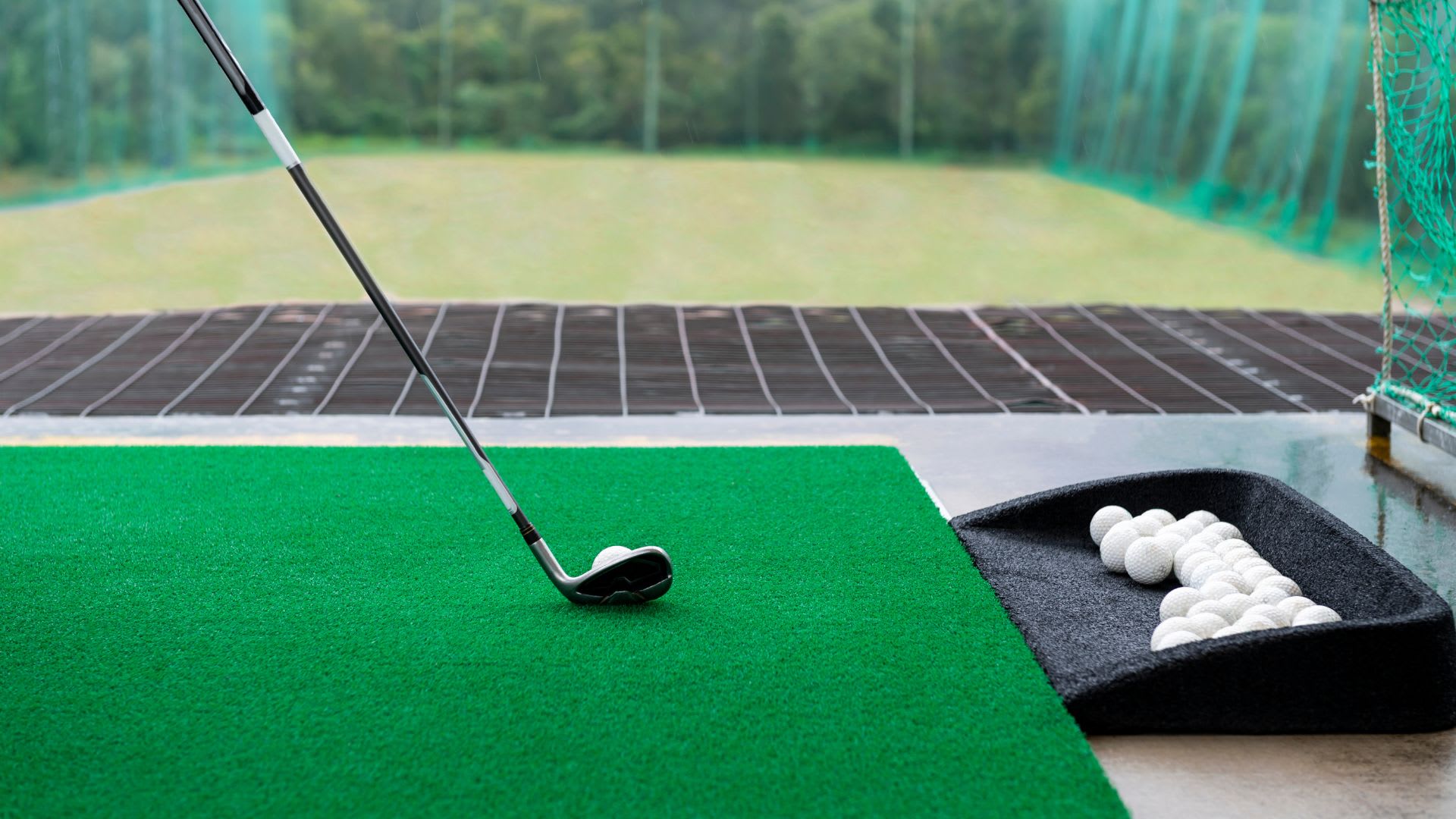 A golf club poised to strike a ball on a green practice mat at a driving range, with a tray of golf balls nearby and a lush green field in the background.